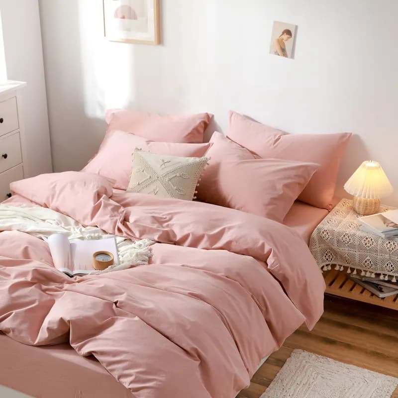 A dusty-pink washed cotton bedding set on a bed with matching pillowcases, a cream tasselled cushion, an open magazine, a rattan table lamp and sunlight across a wooden floor