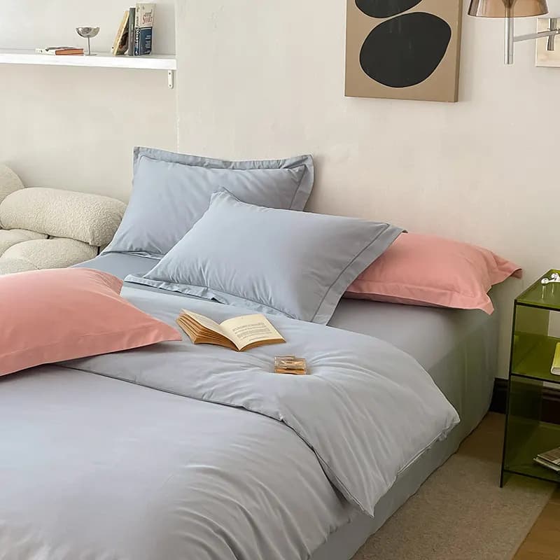 A grey-blue and dusty-pink two-tone reversible bedding set on a bed with matching pillowcases, an open book resting on the duvet, a green glass side table and an abstract print above