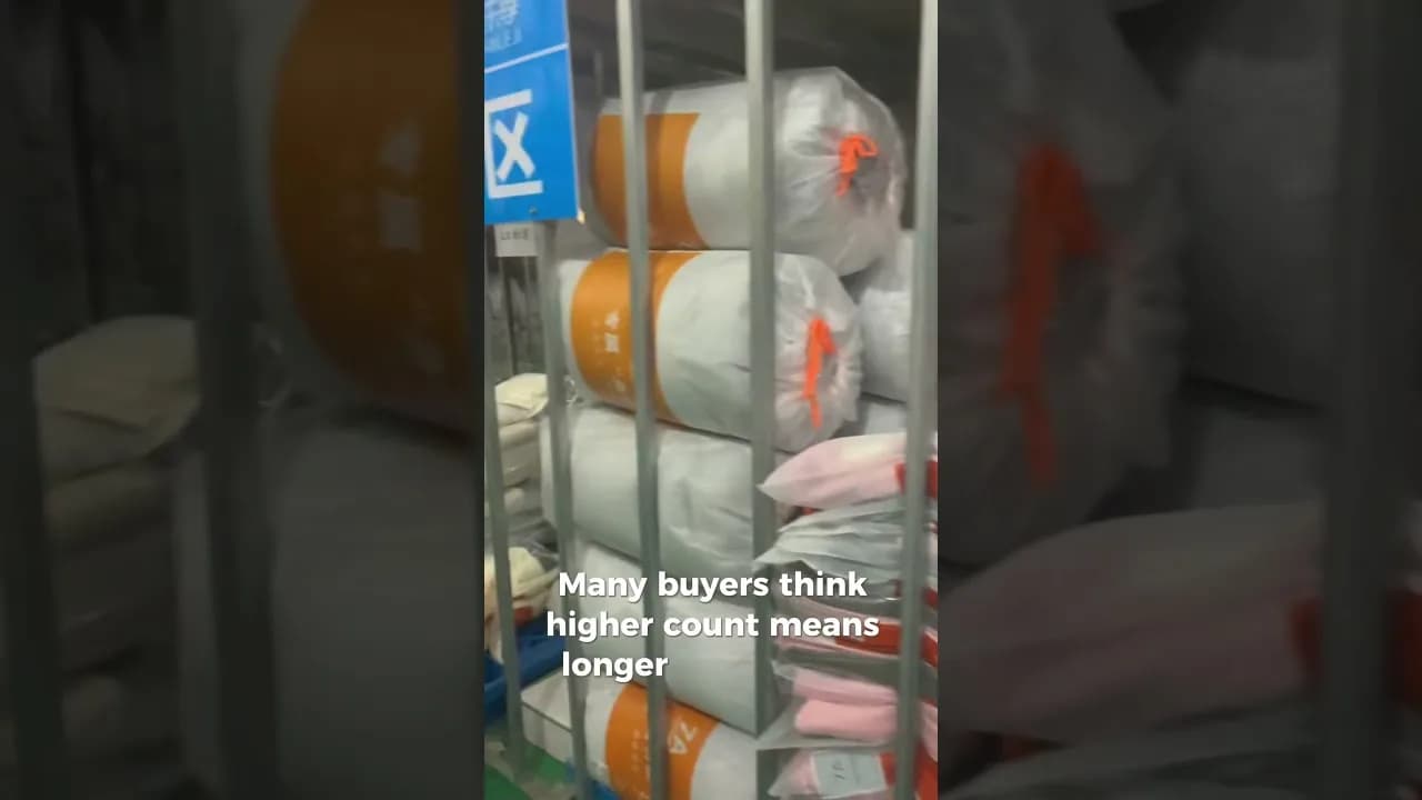 Rolls of plastic-wrapped white cotton fabric bolts with orange labels stacked in a metal warehouse rack at a textile mill, with an on-screen caption noting that buyers assume a higher count lasts longer.