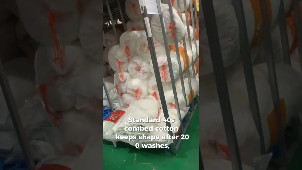 Plastic-wrapped bundles of white combed cotton tied with orange cord, stacked on a metal warehouse rack at a textile mill, with an on-screen caption about combed cotton keeping its shape after 200 washes.