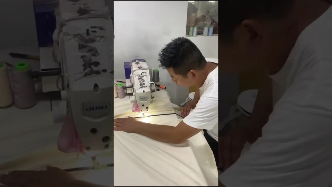 Video frame showing an operator guiding a large light-coloured bedding panel through an industrial sewing machine