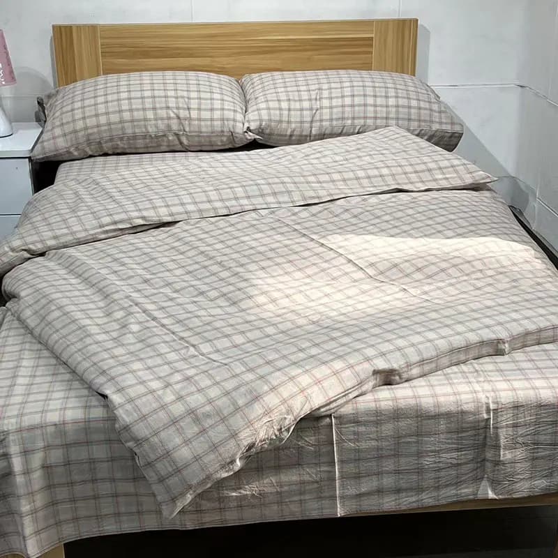 A beige and pink fine-check cotton bedding set on a bed with a light wood headboard in a plain, practical room, with matching pillowcases and a white nightstand