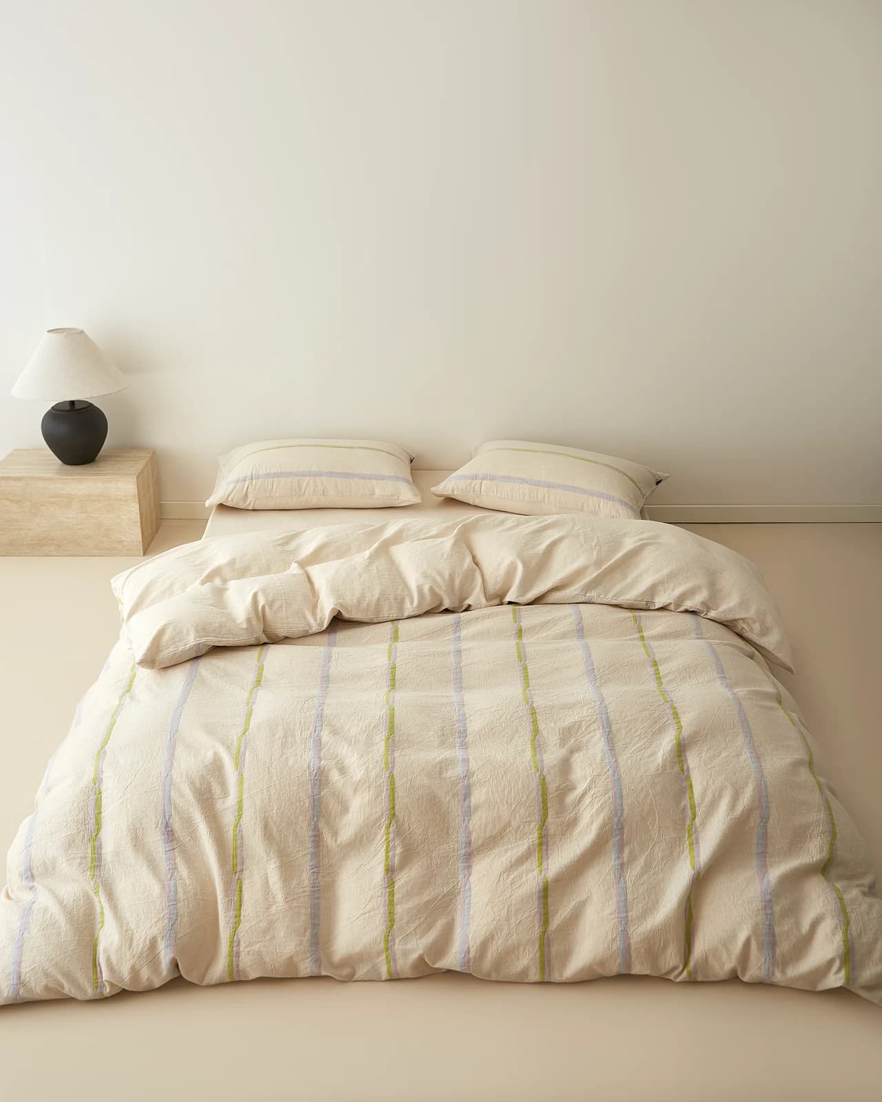 Cream washed-cotton duvet set with narrow vertical lilac and lime-green woven stripes on a low bed, two matching pillows, travertine nightstand and black ceramic lamp in a bare beige room