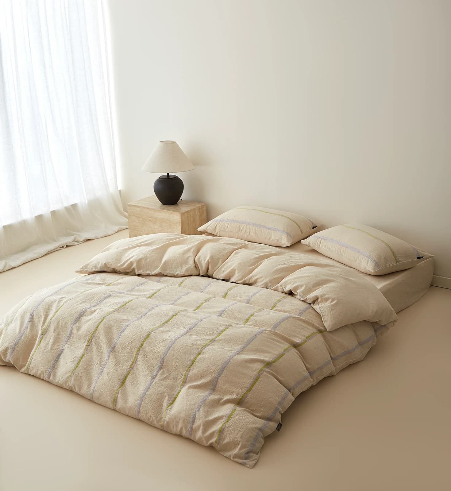 A natural beige washed-cotton set with fine lilac and lime-green pinstripes on a low floor-level bed, sheer white curtains behind and a black round lamp on a travertine cube