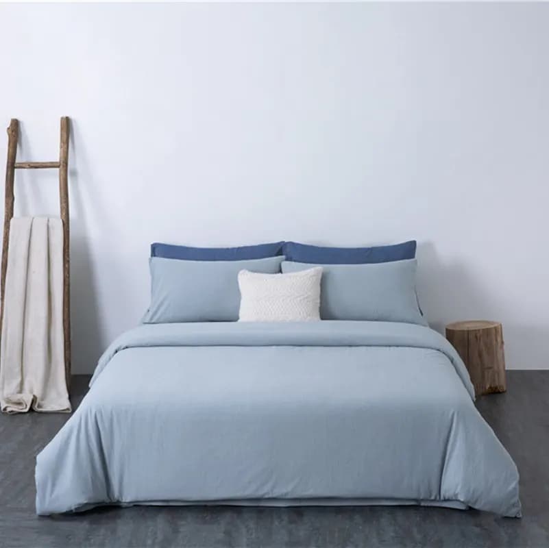Pale blue solid cotton duvet set with navy pillowcases and a white cable-knit cushion on a bed, beside a weathered wooden ladder draped with a cream throw and a tree-stump stool
